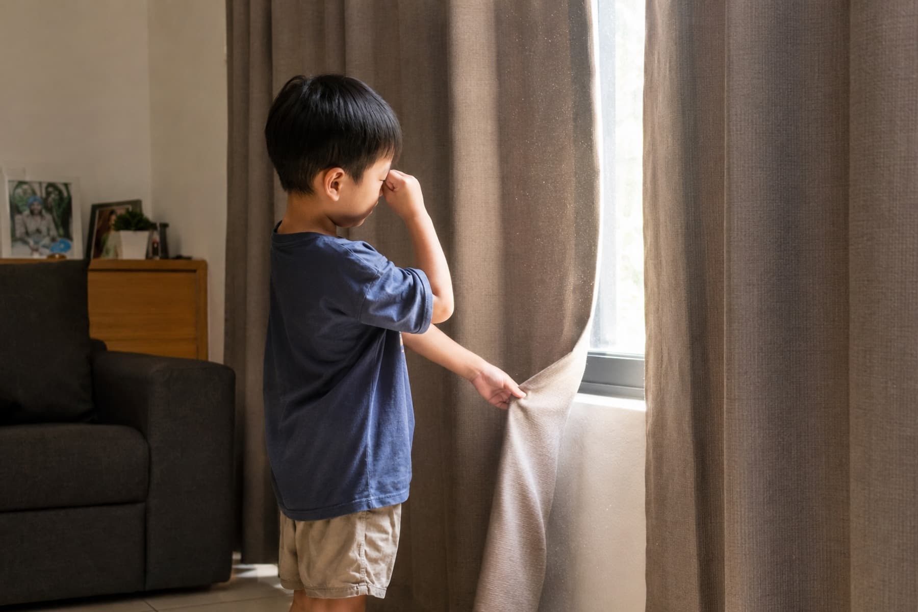 Child rubbing his eye while pulling back a curtain, dust visible in the window light