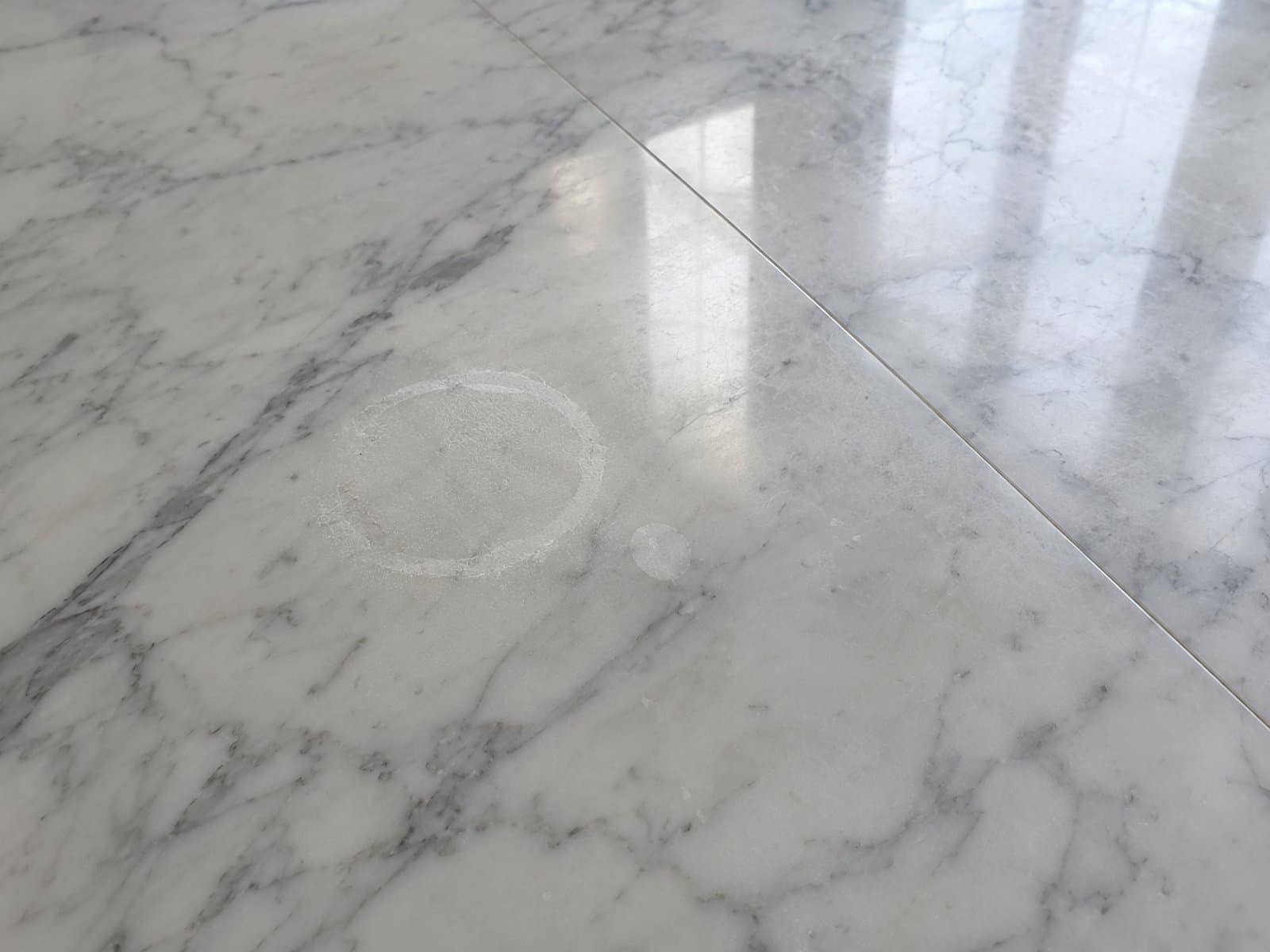 Pale dull ring and a smaller round mark on grey-veined marble beside reflected window light