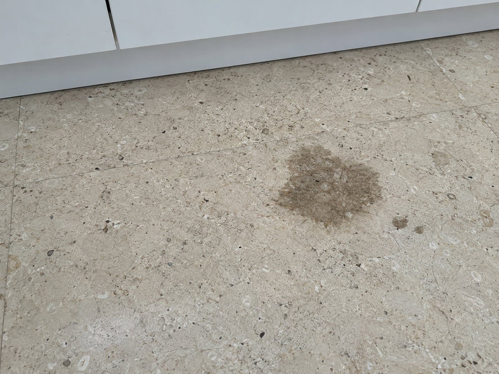 Irregular dark patch with smaller dark spots on beige stone flooring beside white kitchen cabinets