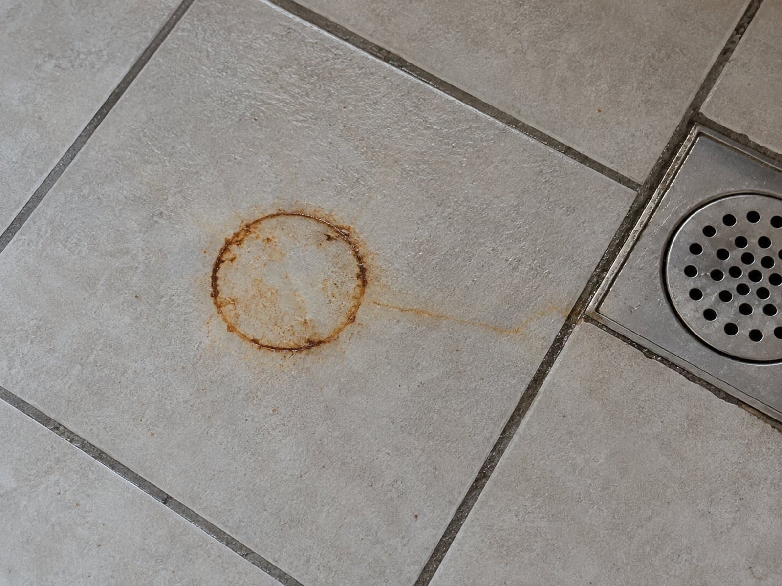 Orange-brown ring with a thin streak on grey bathroom tile beside a metal floor drain