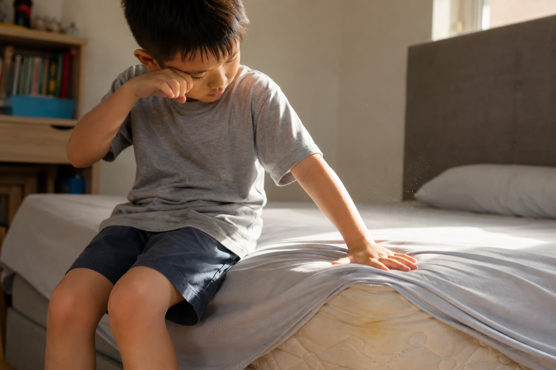 Child rubbing his eye while dust rises from the mattress where his hand presses, a faint stain visible on the exposed corner