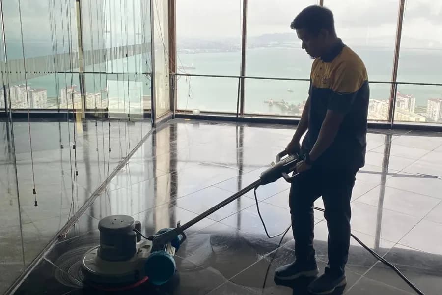 Yellowbee technician in a yellow and navy uniform polishing dark glossy floor tiles with a rotary machine beside large windows