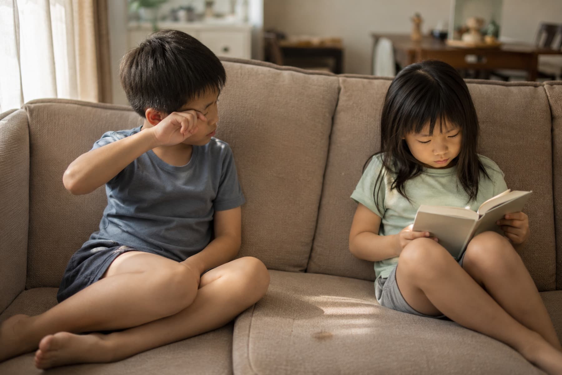Child rubbing his eye while sitting on a stained sofa cushion next to his sister